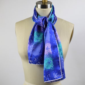 May include: A blue and purple silk scarf with a white border and a white signature that says "Usna" on the bottom right corner. The scarf is tied around a dress form's neck.