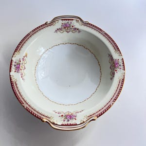 May include: Cream-colored, round serving bowl with a burgundy and gold rim. The rim features a repeating pattern and floral accents. The bowl has two small handles and a floral design in the center.