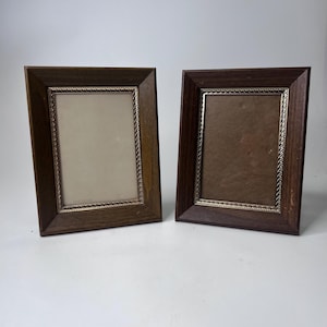 May include: Two rectangular picture frames with dark brown wooden borders and silver-colored inner trim. The frames are empty, with one having a light-colored backing and the other a darker backing. The frames are displayed against a white background.