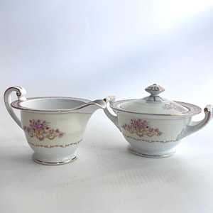 Può includere: Servizio di lattiera e zuccheriera con coperchio in porcellana color crema. Entrambi i pezzi presentano un motivo floreale con fiori rosa e viola, finiture dorate e un delicato bordo rosa. La lattiera ha un manico e un beccuccio.