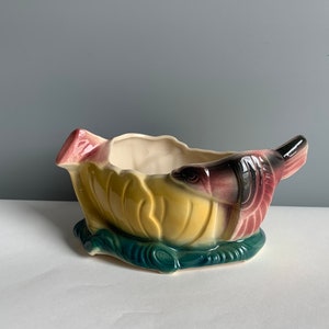 Może przedstawiać: Ceramiczna doniczka w kształcie ryby pływającej w lilii wodnej. Doniczka jest pomalowana w odcieniach zieleni, żółci, różu i brązu.