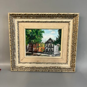 May include: Framed painting of a street scene with three houses, two trees, and a blue sky. The houses are painted in shades of red, gray, and white. The trees are green and the sky is blue with white clouds. The painting is signed "Ian Selmas" in the lower right corner.