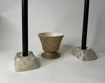 May include: Two ceramic candle holders with a textured design, each holding a black candle. A matching ceramic bowl with a textured design is in the center.