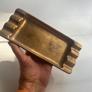 Op de afbeelding: Een rechthoekige asbak in antieke stijl met een golvende rand. De asbak is gemaakt van een bronskleurig metaal en heeft een licht getextureerd oppervlak. Het heeft een klassiek ontwerp met groeven om sigaretten op te laten rusten.