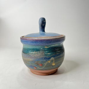 May include: A small, blue and green ceramic jar with a lid and a handle. The jar has a swirl pattern and red and yellow accents.