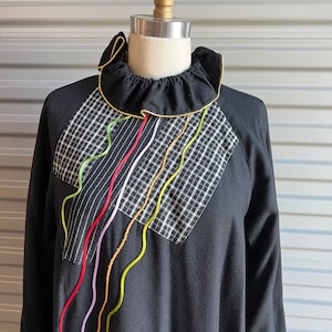 May include: Black long-sleeved shirt with a ruffled collar and a plaid fabric panel on the front. The panel has colorful, wavy lines sewn on top.