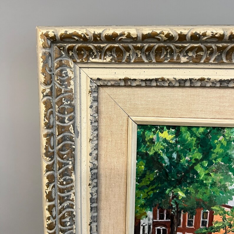 May include: A framed painting of a street scene with a large green tree in the foreground. The frame is a light beige color with a gold-colored decorative border.