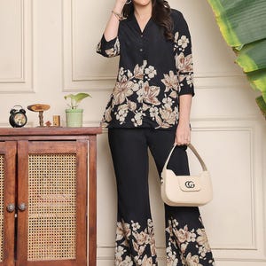 May include: Black floral print top and pants set. The top has a v-neck and three-quarter sleeves. The pants are flared at the bottom. A cream-colored handbag with a gold-colored clasp is held in the hand. Black sandals complete the outfit.