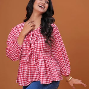 May include: A red and white gingham print top with long sleeves and a tie detail at the neckline. The top has a peplum hem and is paired with blue jeans. A gold bracelet is visible on the wrist.