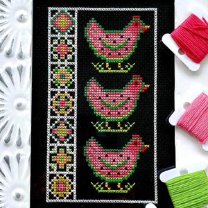 May include: Cross-stitch embroidery showing three pink chickens with green detailing on a black backdrop. A decorative border with geometric patterns is present. Spools of pink and green thread are also visible.