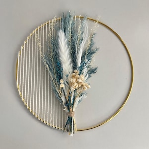 May include: A circular wall hanging featuring a gold metal ring with a woven white yarn design on the left side. A bundle of dried flowers in shades of blue, white, and beige is tied to the ring with twine. The wall decor is a modern, minimalist design.
