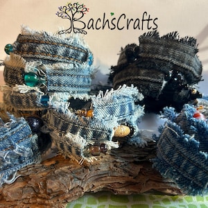 Puede incluir: Cinco pulseras de mezclilla con bordes deshilachados y detalles de cuentas descansan sobre un trozo de madera. Las pulseras son de varios tonos de azul y negro. La imagen incluye el texto "Bach's Crafts".