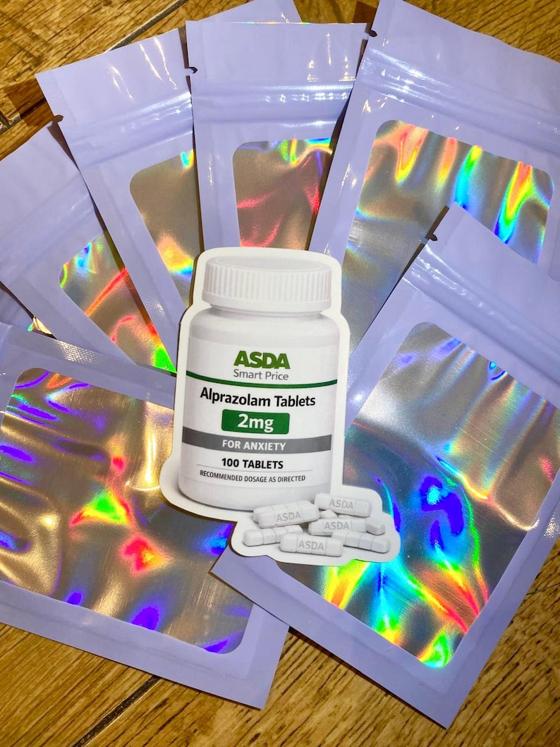 Op de afbeelding: Een productfoto met een witte ASDA Alprazolam Tabletten fles en verschillende verzegelde, iriserende zakjes. Op het flesetiket staat "2mg" en "VOOR ANGST, 100 TABLETTEN". Er zijn meerdere tabletten zichtbaar. De zakjes hebben een paarse rand.