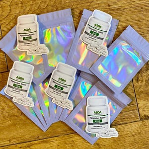 Op de afbeelding: Een assortiment stickers met witte pillenflessen met het opschrift "ASDA Alprazolam Tablets 2mg" en "100 TABLETS" met een kleine stapel pillen. De stickers zijn geplaatst op iriserende, holografische zakjes.