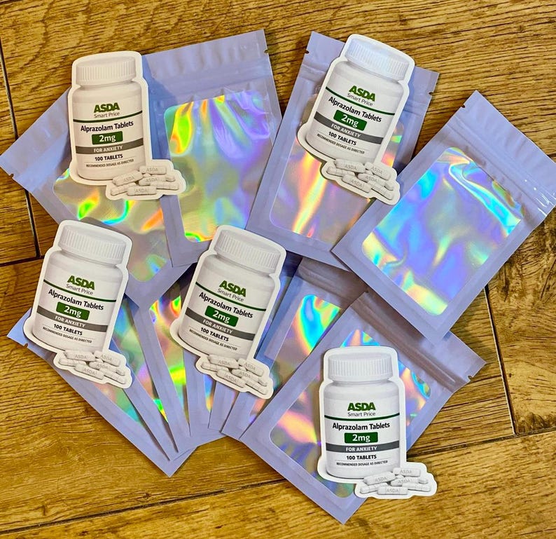 Op de afbeelding: Een assortiment stickers met witte pillenflessen met het opschrift "ASDA Alprazolam Tablets 2mg" en "100 TABLETS" met een kleine stapel pillen. De stickers zijn geplaatst op iriserende, holografische zakjes.