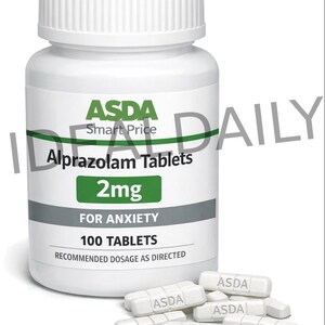 Op de afbeelding: Een witte plastic fles ASDA Alprazolam Tabletten, 2mg, voor angst. Het etiket geeft 100 tabletten en de aanbevolen dosering aan. Verschillende witte pillen met "ASDA" zijn verspreid in de buurt van de fles.