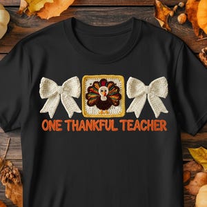 May include: Black t-shirt with the text "ONE THANKFUL TEACHER" in orange. The shirt features a crochet design of a turkey in a square frame, flanked by two white crochet bows. Pumpkins and fall leaves surround the shirt.