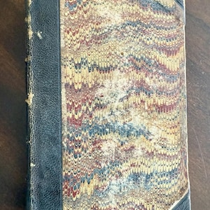 May include: An antique book featuring a marbled cover in yellow, red, and blue hues. The spine and corners are bound in dark blue leather. The book exhibits signs of age and wear, with a distressed appearance.