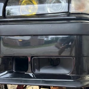 Puede incluir: Primer plano de un parachoques de coche negro con un acabado brillante. El parachoques tiene dos aberturas rectangulares y una rejilla de malla. Los faros están visibles arriba, con un tinte amarillo en las luces interiores.