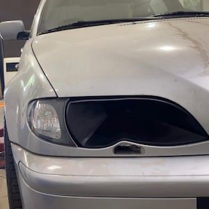 May include: Silver car with a black headlight and a black front bumper. The vehicle is parked in a garage or workshop. The image is a close-up of the front of the car.