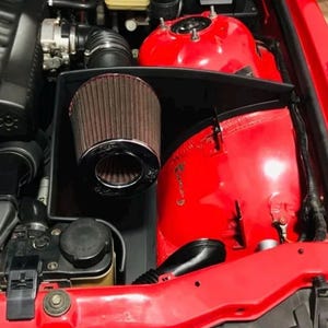 Può includere: Vano motore con filtro dell'aria marrone e componenti rossi. Il filtro dell'aria ha un bordo cromato ed è posizionato all'interno di un alloggiamento nero. Le parti del motore circostanti sono nere e rosse, con vari tubi e raccordi visibili. La scena suggerisce manutenzione o modifica automobilistica.