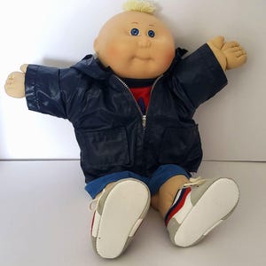 May include: A vintage Cabbage Patch Kid doll with a bald head and blue eyes. The doll is wearing a navy blue jacket, red shirt, blue shorts, and gray and white sneakers. The doll's arms are outstretched.