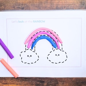 Peut inclure: Un arc-en-ciel coloré en pâte à modeler rose, violet et bleu, avec deux nuages souriants dessinés. Le texte "Let's look at the RAINBOW" est au-dessus de l'arc-en-ciel. Deux morceaux de pâte à modeler, violet et orange, sont sur la surface en bois.