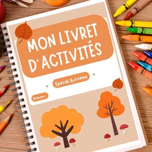May include: A spiral-bound activity book with the text "MON LIVRET D'ACTIVITÉS" and "Spécial Automne" in orange. The cover features illustrations of autumn trees and mushrooms. Crayons and paint supplies surround the book on a wooden surface.