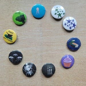 Op de afbeelding: Een verzameling ronde buttons met diverse ontwerpen. De buttons tonen illustraties van een kwal, sterren, een cupcake, een kat en andere afbeeldingen. Sommige buttons hebben tekst, waaronder "gnarp" en "Oops! She treaded."