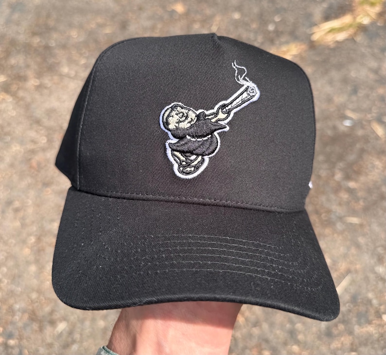Pode incluir: Bon&eacute; de beisebol preto com aba reta. O bon&eacute; apresenta um patch bordado de um personagem de desenho animado fumando um cigarro enrolado. A figura est&aacute; vestindo uma t&uacute;nica. O bon&eacute; &eacute; feito de tecido dur&aacute;vel.