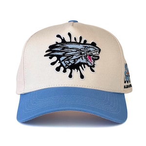 Pode incluir: Boné de beisebol com coroa bege clara e aba azul clara. O boné apresenta um design bordado de cabeça de dragão com efeito de respingo preto. O dragão tem detalhes azuis e brancos com boca rosa.