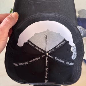 Pode incluir: Bon&eacute; de beisebol preto com aba curva. A parte inferior &eacute; branca com um desenho preto e o texto "HeadHunters" em branco. O bon&eacute; &eacute; segurado por uma m&atilde;o.