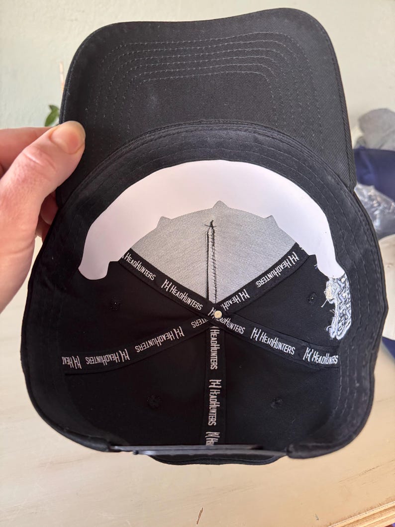 Pode incluir: Bon&eacute; de beisebol preto com aba curva. A parte inferior &eacute; branca com um desenho preto e o texto "HeadHunters" em branco. O bon&eacute; &eacute; segurado por uma m&atilde;o.