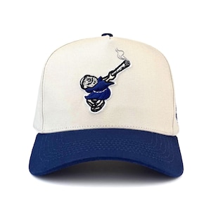 Puede incluir: Gorra de béisbol con corona color crema y visera azul marino. La gorra presenta un parche bordado de un personaje de dibujos animados haciendo un dab mientras sostiene un cigarrillo encendido. El personaje lleva un atuendo azul.