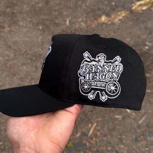 Pode incluir: Bon&eacute; de beisebol preto com aba reta. O bon&eacute; apresenta texto bordado branco e um gr&aacute;fico de uma carro&ccedil;a com as palavras "Banned Wagon" na lateral. O bon&eacute; &eacute; segurado por uma m&atilde;o.