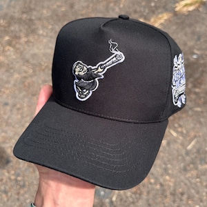 Pode incluir: Bon&eacute; de beisebol preto com aba reta. O bon&eacute; apresenta um patch bordado de um personagem de desenho animado fumando um cigarro. Um segundo patch com texto &eacute; vis&iacute;vel na lateral.