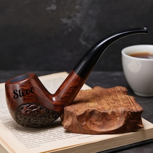 May include: A wooden tobacco pipe with a curved black stem and a brown bowl. The name "Steve" is engraved on the bowl. The pipe rests on a wooden block, with a cup of coffee in the background.