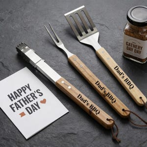 Puede incluir: Un juego de utensilios para barbacoa con mangos de madera, que incluye pinzas, un tenedor y una espátula, cada uno grabado con "Dad's BBQ". También hay un pequeño frasco de condimentos y una tarjeta que dice "Happy Father's Day".