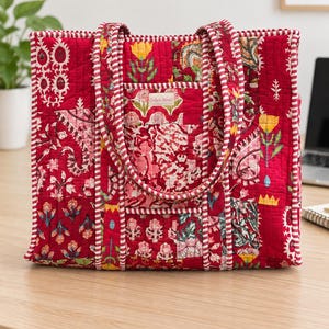 May include: A quilted tote bag in a vibrant red fabric featuring a multi-colored floral and paisley block print pattern. The bag has two matching striped handles and a small rectangular label on the front that reads Color Nest in script.