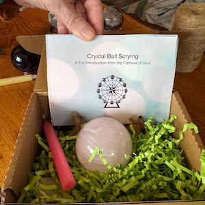 May include: A rose quartz crystal ball rests in a cardboard box filled with green paper shreds. A pink candle and a booklet titled "Crystal Ball Scrying" are also in the box. The booklet features a Ferris wheel design.