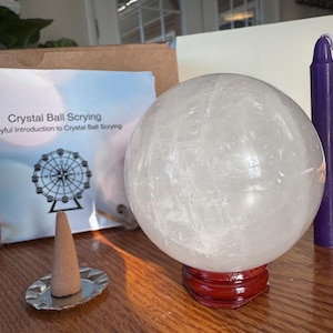 May include: A clear crystal ball on a red wooden stand, with a purple candle and a booklet titled "Crystal Ball Scrying." An incense cone sits on a silver dish. The booklet has a Ferris wheel illustration.