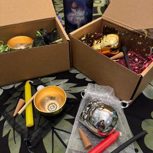 May include: Two open cardboard boxes containing spiritual items. One box holds a gold bowl, a yellow candle, and incense cones. The other box contains a gold decorative item and a deck of cards titled "Shaman's Dream". Additional items are displayed on a patterned surface.