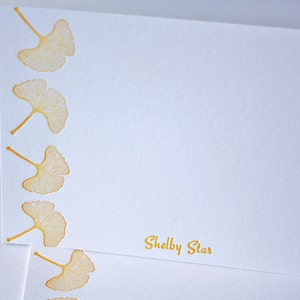 May include: White stationery card with a gold border and a gold ginkgo leaf design. The card has the text "Shelby Star" printed in gold at the bottom.