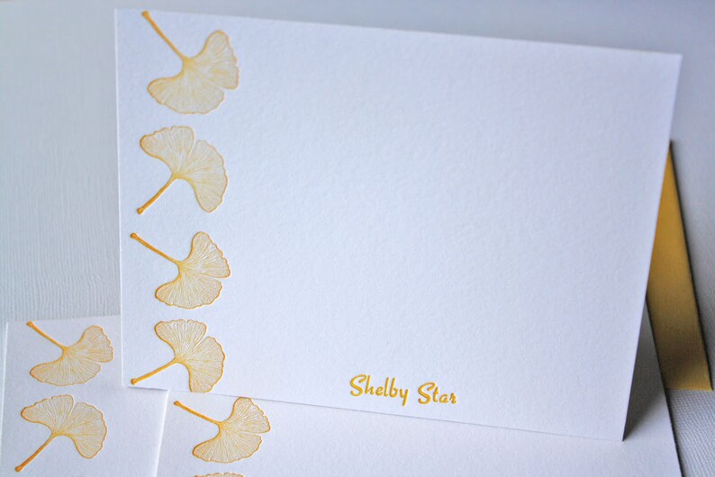 May include: White stationery card with a gold border and a gold ginkgo leaf design. The card has the text "Shelby Star" printed in gold at the bottom.
