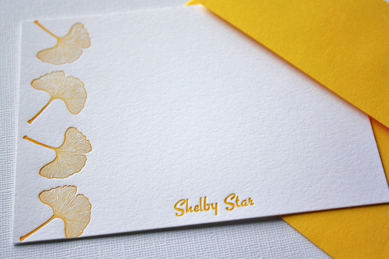 May include: White stationery card with a yellow envelope. The card has a border of five gold ginkgo leaves and the text "Shelby Star" in gold lettering.