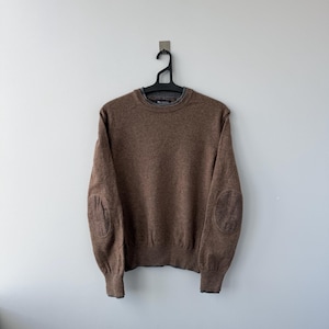 Puede incluir: Un suéter marrón con coderas, colgado de una percha negra contra una pared blanca. El suéter tiene cuello redondo y puños acanalados. Las coderas son de una tela marrón más oscura con estampado.