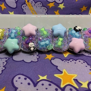 May include: A set of six artificial nails, each adorned with glitter, star-shaped charms, and miniature decorative elements. The nails feature a variety of colors, including pink, blue, and green, with small panda and skeleton designs.
