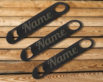 May include: Three black bottle openers with the word "Name" in a stylized font. The openers are arranged diagonally on a wooden surface. Each opener has a circular hole at one end.