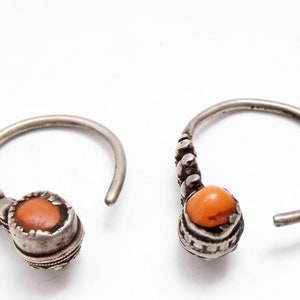 May include: A pair of silver hoop earrings featuring a textured, oxidized metal finish and a single round, orange-colored stone set in a bezel at the base of each hoop. The hoops have a curved wire design with small decorative metal beads.