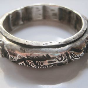 May include: A silver-toned metal bangle bracelet featuring a raised, textured relief pattern of stylized fish or aquatic creatures wrapping around the exterior surface. The bracelet has a thick, rounded profile with a polished finish and dark recessed areas.